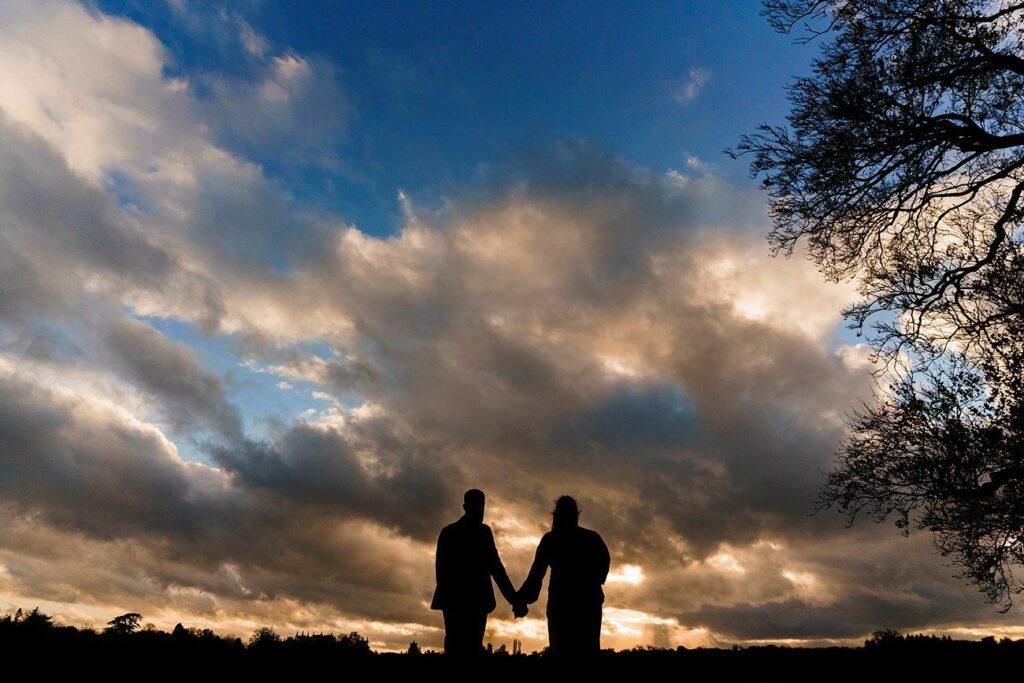 Two people hold hands while standing outdoors against a dramatic backdrop of a sunset sky filled with large, dark clouds. Silhouetted trees frame the scene on either side, creating a serene and romantic atmosphere. Image by Documentary Wedding Photographer Lewis Bishop Photography.