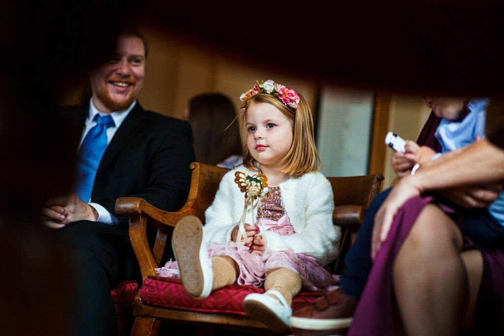 A young girl sits on a chair wearing a white cardigan, pink dress, and a floral headband, holding a toy. A smiling man in a suit is next to her. The background is slightly blurred with other people partially visible, capturing a warm, indoor setting. Image by Documentary Wedding Photographer Lewis Bishop Photography.