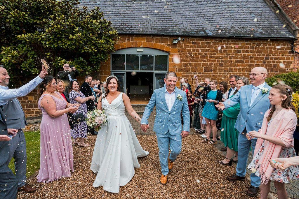 A newlywed couple walks down an aisle outdoors with guests throwing confetti, as they smile holding hands, with people clapping and taking photos in the background. Image by Documentary Wedding Photographer Lewis Bishop Photography.