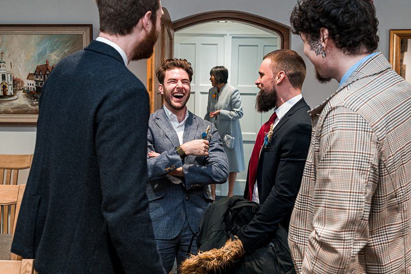 Four men in suits are standing and talking indoors. One man is laughing heartily while the others are smiling and engaged in conversation. A woman is visible in the background through a doorway. Image by Documentary Wedding Photographer Lewis Bishop Photography.