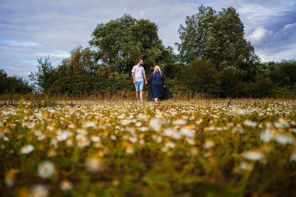 A couple walks hand in hand through a field of wildflowers. The woman on the right is dressed in a dark navy dress, and the man on the left wears a white T-shirt and shorts. Behind them, there are tall, leafy trees and bushes under a partly cloudy sky. Image by Documentary Wedding Photographer Lewis Bishop Photography.