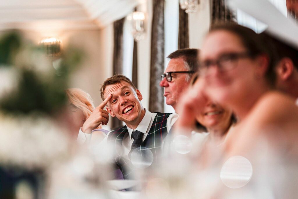 A man in a checkered vest and tie smiles broadly as he points to his cheek, seated at a formal event with others who are blurred in the foreground and background. The setting appears festive, with bright lighting and elegant decor. Image by Documentary Wedding Photographer Lewis Bishop Photography.