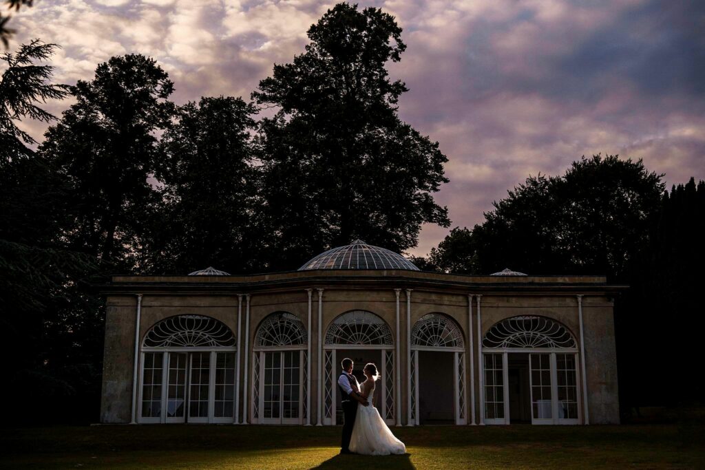 A couple dressed in wedding attire stands close together in front of an elegant building with tall windows, surrounded by trees. The sky above them is filled with dramatic clouds, creating a romantic and atmospheric scene. Image by Documentary Wedding Photographer Lewis Bishop Photography.
