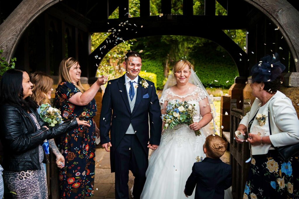 A bride and groom, dressed in traditional wedding attire, are showered with confetti by their smiling guests under a wooden archway. The bride holds a bouquet, and a young child, facing away, is seen at the front, adding to the joyful moment. Image by Documentary Wedding Photographer Lewis Bishop Photography.
