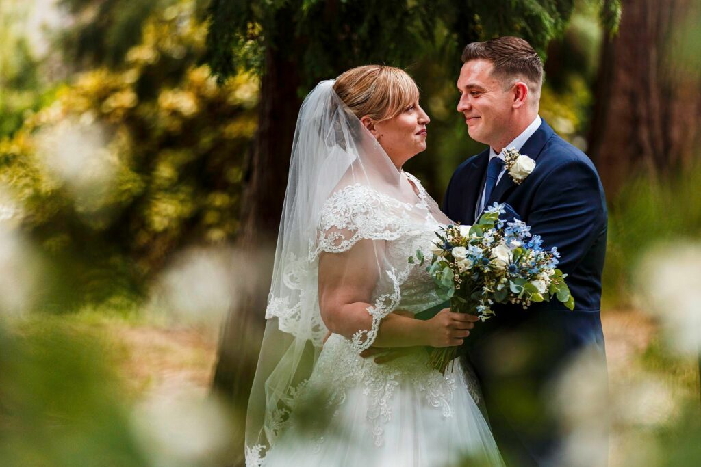 A bride and groom are standing close together in a lush, green outdoor setting. The bride is wearing a white lace wedding dress and veil, holding a bouquet of white and blue flowers. The groom is wearing a dark blue suit with a white boutonniere, smiling at each other lovingly. Image by Documentary Wedding Photographer Lewis Bishop Photography.