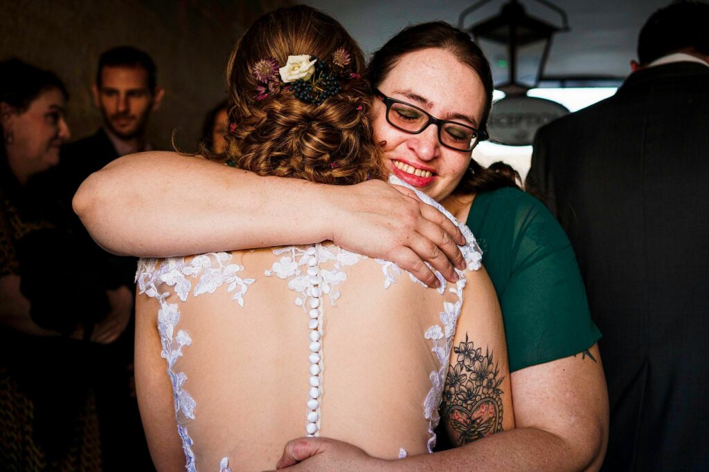 A woman in a white dress with floral lace details and an intricate updo is hugging another woman wearing glasses and a green dress. The woman in glasses is smiling warmly. A group of people is visible in the background, partially out of focus. Image by Documentary Wedding Photographer Lewis Bishop Photography.