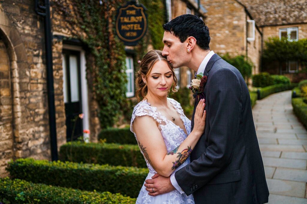 A groom kisses the forehead of his bride as they stand embracing outside a stone building with a sign reading "The Talbot Hotel." The bride wears a lace wedding dress and the groom is in a suit. The pathway and greenery in the background add to the romantic setting. Image by Documentary Wedding Photographer Lewis Bishop Photography.