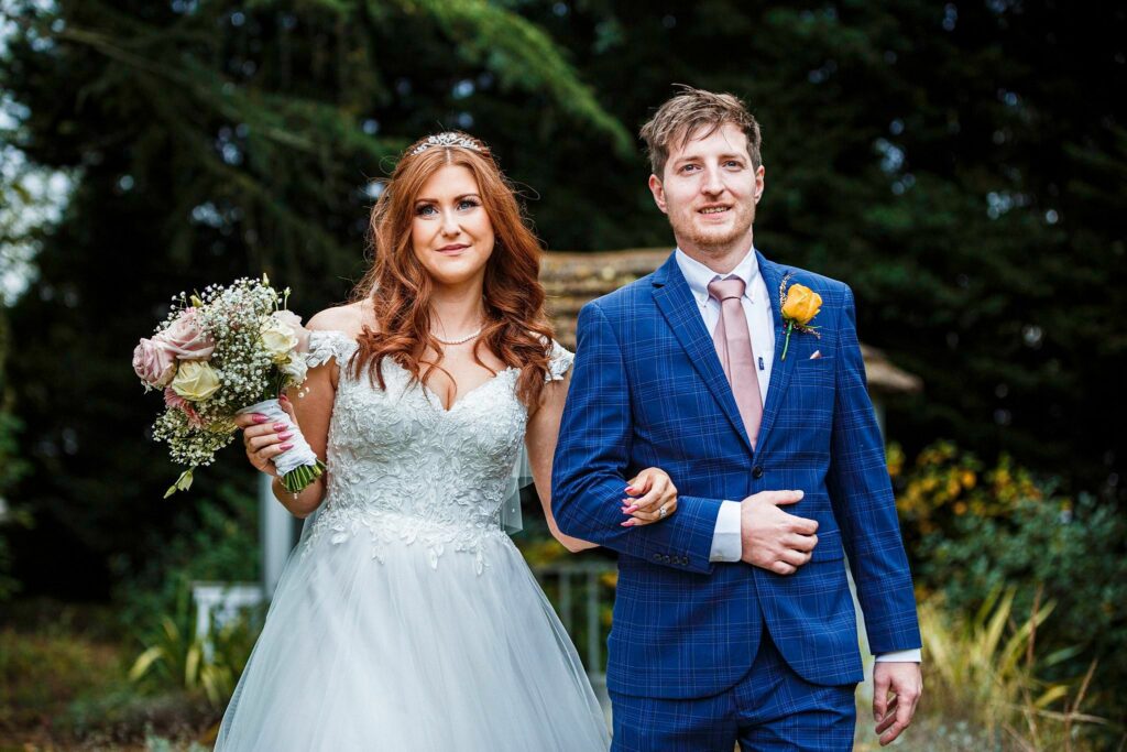 A bride and groom walk outdoors, the bride holding a bouquet of flowers and wearing a white wedding dress and tiara, while the groom wears a blue checkered suit and a pink tie. They appear happy, with greenery and a gazebo visible in the background. Image by Documentary Wedding Photographer Lewis Bishop Photography.