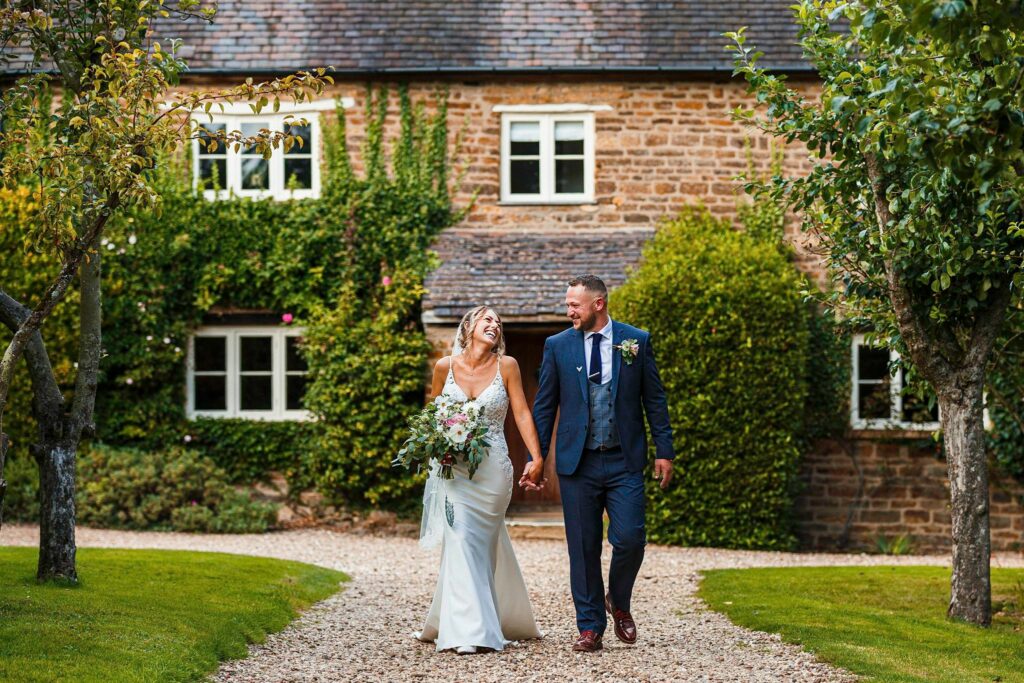 A smiling couple in wedding attire walk hand in hand on a gravel pathway. The bride, wearing a white dress and holding a bouquet, looks at the groom, who is dressed in a dark suit. They walk towards a stone house covered in green ivy, framed by trees. Image by Documentary Wedding Photographer Lewis Bishop Photography.