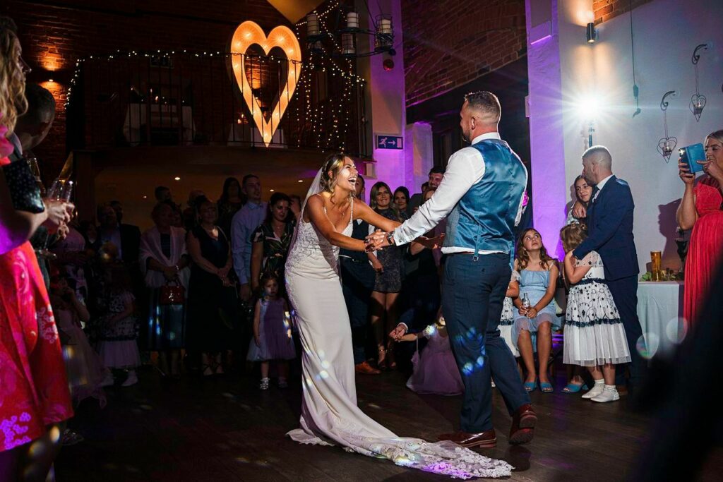 A bride in a white gown and a groom in a vest and tie share their first dance on a dimly lit dance floor surrounded by guests. A large, illuminated heart decoration and string lights are visible in the background, creating a romantic atmosphere. Image by Documentary Wedding Photographer Lewis Bishop Photography.