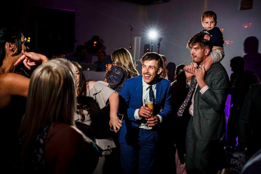 A lively group of people are dancing at a party. One man in a blue suit smiles while holding a drink, and another man with a child on his shoulders laughs. The room is dimly lit with purple and white lights, and various people are seen enjoying themselves in the background. Image by Documentary Wedding Photographer Lewis Bishop Photography.