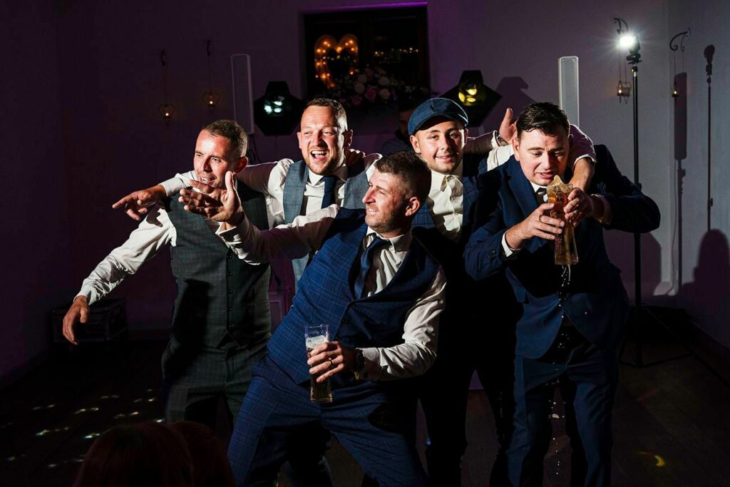 Five men in formal attire are joyfully posing and laughing together at an indoor event. One holds a drink, and another has an arm around his friend's shoulder. The atmosphere is festive, with colorful lights in the background hinting at a party or celebration. Image by Documentary Wedding Photographer Lewis Bishop Photography.