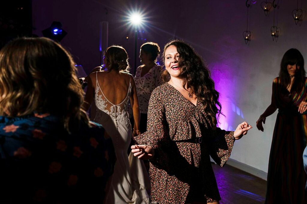 A woman with long, curly hair and a floral dress dances joyfully in a dimly lit room with other people. One person in the background wears a white, patterned dress and another wears a striped dress. A bright light shines from a fixture behind them. Image by Documentary Wedding Photographer Lewis Bishop Photography.