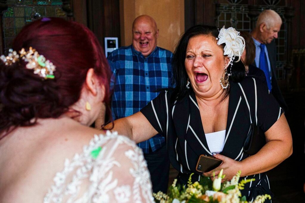 A woman with a white floral accessory in her hair reacts joyfully, holding a phone, as a bride in a white lace dress faces her. A man in a blue plaid shirt is laughing in the background along with another man in a suit. Image by Documentary Wedding Photographer Lewis Bishop Photography.