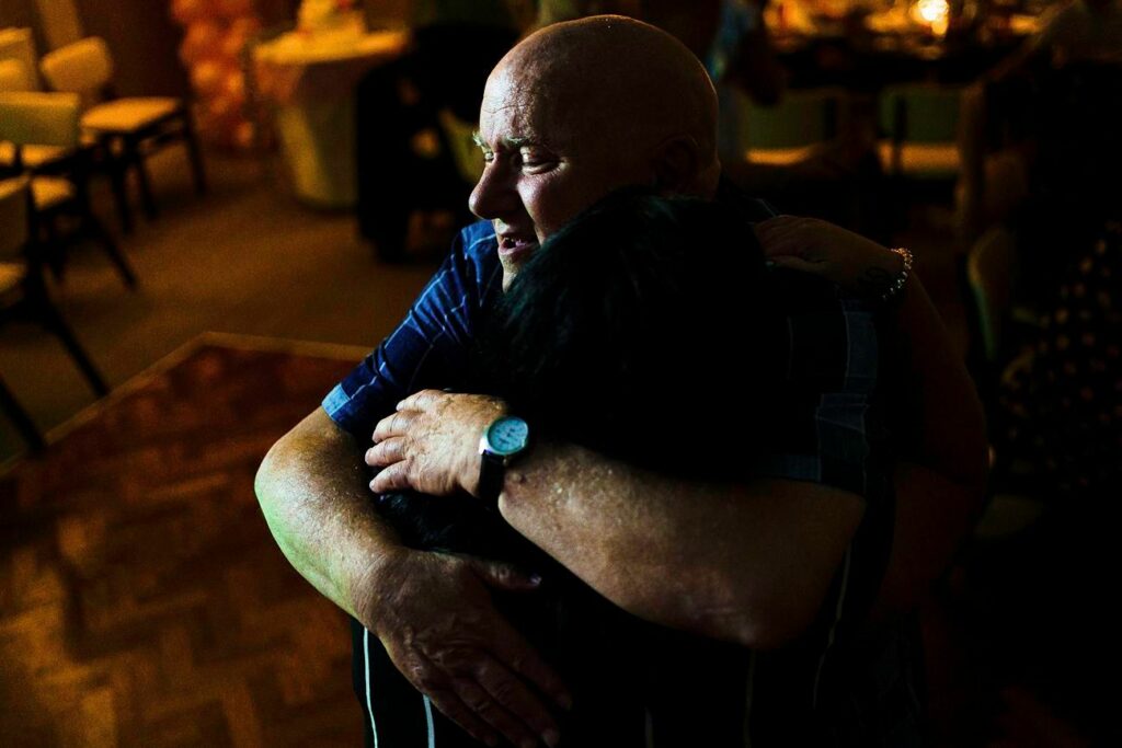 A bald man wearing a striped shirt and wristwatch embraces another person in a dimly lit room, with soft, warm lighting in the background creating an intimate and emotional atmosphere. Image by Documentary Wedding Photographer Lewis Bishop Photography.