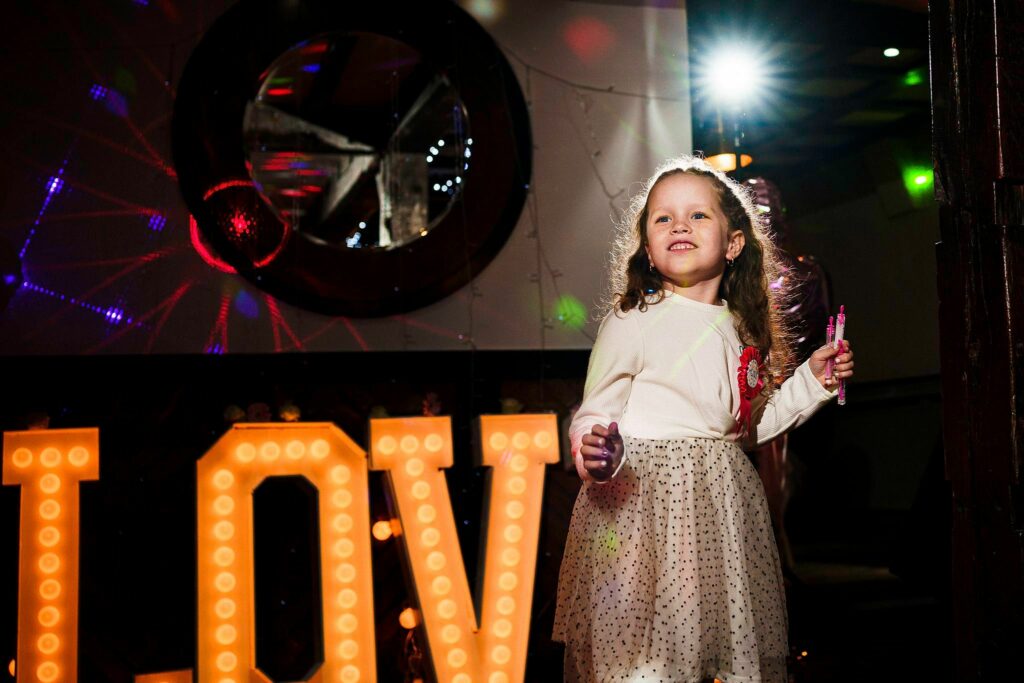 A young girl in a white dress with polka dots stands in front of illuminated letters spelling "LOVE." She has light brown hair and is smiling, holding a small object in her hand. The background features colorful lights and a circular mirror. Image by Documentary Wedding Photographer Lewis Bishop Photography.