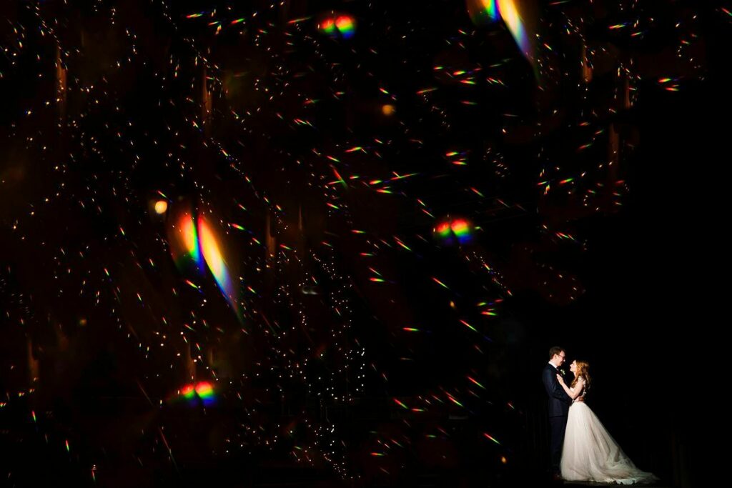 A bride and groom share a romantic moment in the lower right corner against a dark background. Colorful, scattered light reflections fill the remaining space, creating a magical, starry ambiance. The bride wears a white dress, while the groom is in a dark suit. Image by Documentary Wedding Photographer Lewis Bishop Photography.