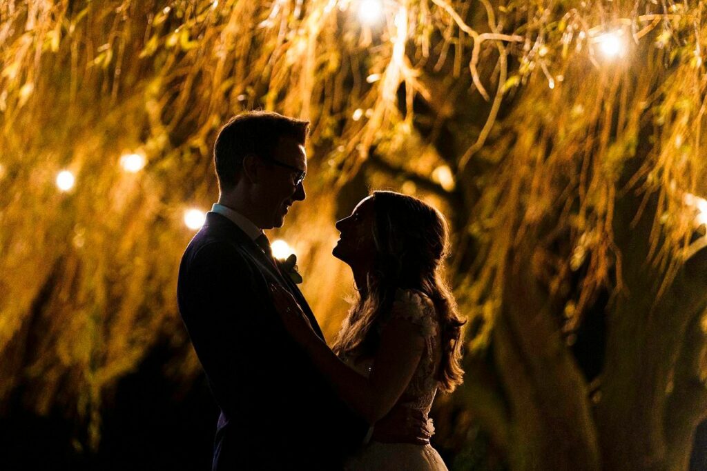 A couple dressed in formal attire share a romantic moment, silhouetted against a background of glowing string lights hanging from tree branches. They are facing each other and appear to be about to kiss, creating a dreamy and intimate atmosphere. Image by Documentary Wedding Photographer Lewis Bishop Photography.