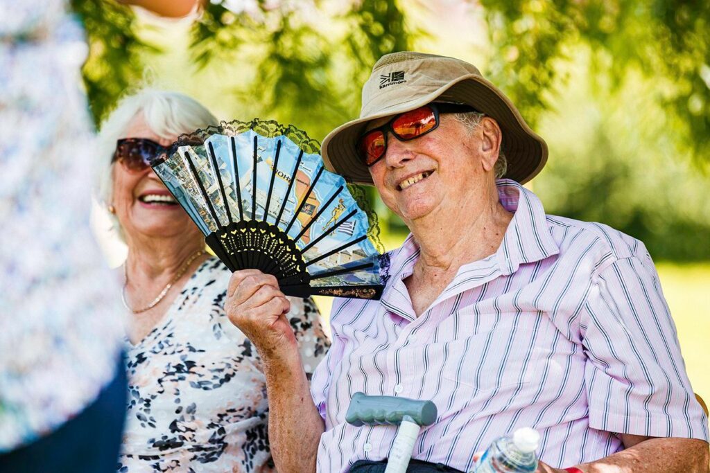Two elderly people are sitting outdoors, smiling and enjoying the day. One person is holding a decorative fan and wearing a hat and sunglasses. The other person, wearing glasses, is dressed in a patterned top. They appear relaxed and happy, with greenery in the background. Image by Documentary Wedding Photographer Lewis Bishop Photography.