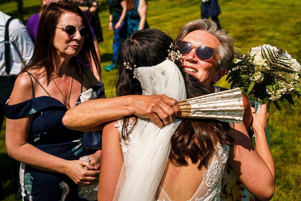 A bride in a white dress and veil hugs an older woman wearing sunglasses while holding a bouquet of flowers. Another woman in a dark off-shoulder dress and sunglasses stands nearby, smiling. They are outdoors on a sunny day, with a grassy field and other people in the background. Image by Documentary Wedding Photographer Lewis Bishop Photography.