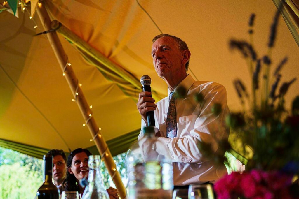 An older man holding a microphone stands under a tent with fairy lights, giving a speech. He is wearing a shirt and tie. Surrounding him, partially visible, are two seated guests listening attentively, with bottles and glasses on the table in front of them. Image by Documentary Wedding Photographer Lewis Bishop Photography.