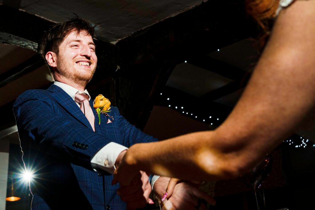 A man in a blue suit with a yellow boutonniere smiles and holds hands with a woman out of frame, engaged in a dance. The background features dim lighting with string lights creating a festive atmosphere. Image by Documentary Wedding Photographer Lewis Bishop Photography.