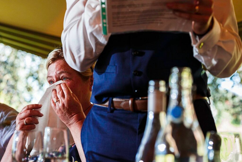A woman wipes tears from her eyes with a napkin, appearing emotional during a speech. The speech is being delivered by a person holding a paper, dressed in a blue waistcoat and white shirt. Various drink bottles are on the table in the foreground. Image by Documentary Wedding Photographer Lewis Bishop Photography.