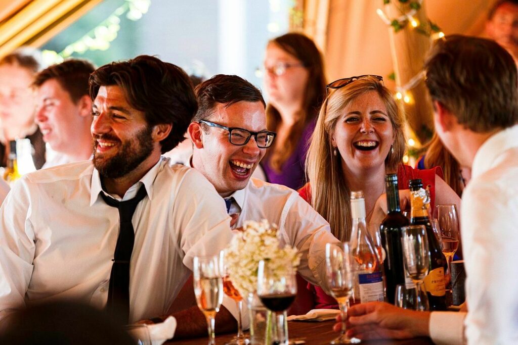 A group of people, dressed in formal attire, are seated at a table and laughing. The table is filled with various bottles and glasses, and there are lights and decorations in the background, suggesting a festive or celebratory event. Image by Documentary Wedding Photographer Lewis Bishop Photography.