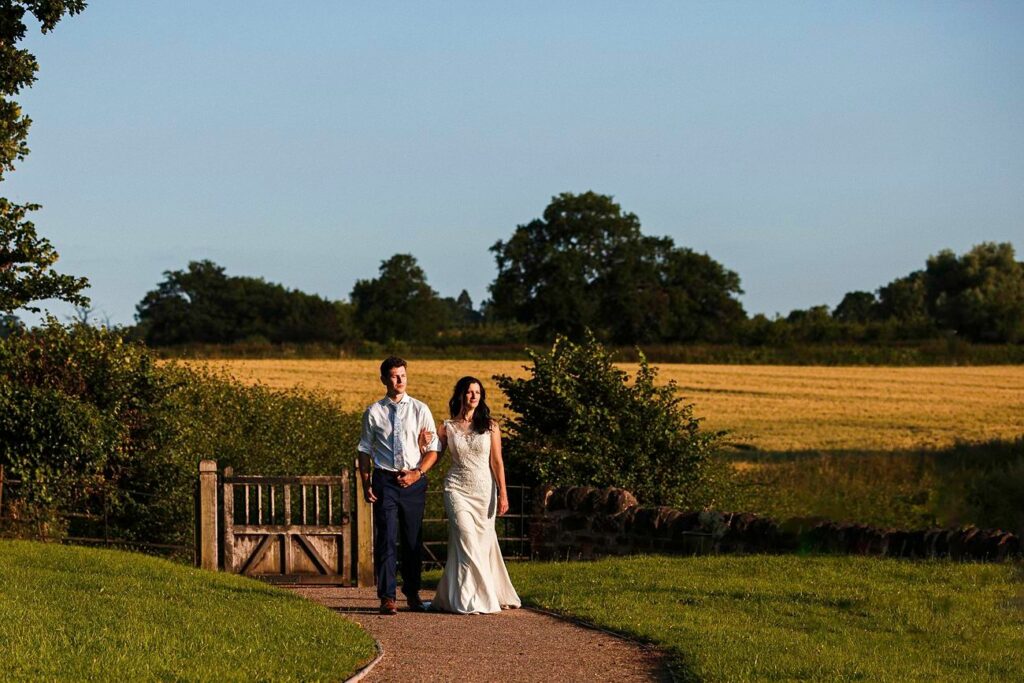 A couple walks hand in hand down a paved path in a scenic countryside setting. The man wears a light blue shirt and dark pants, while the woman wears a white dress. They are surrounded by green grass, trees, and a golden field under a clear blue sky. Image by Documentary Wedding Photographer Lewis Bishop Photography.