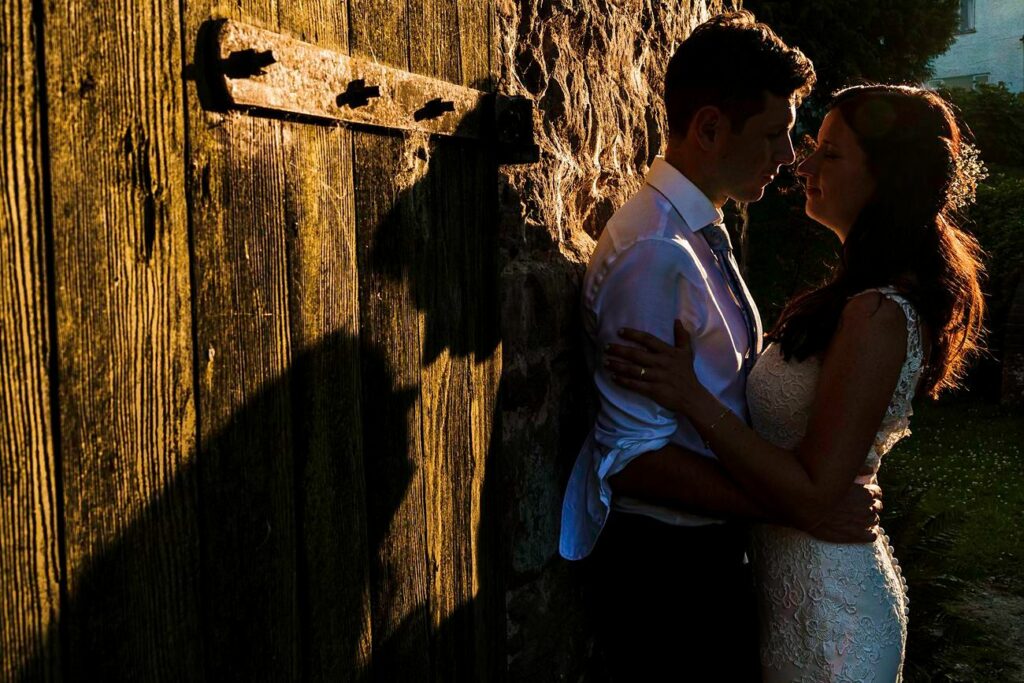 A couple in wedding attire embraces intimately against a rustic wooden door. Their shadows are cast dramatically on the door by the warm glow of the setting sun, creating a romantic and serene atmosphere. The bride's lace gown and the groom's shirt sleeves are visible. Image by Documentary Wedding Photographer Lewis Bishop Photography.
