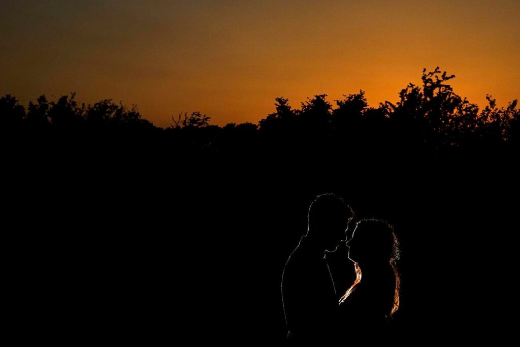 Silhouette of a couple standing close together against a vibrant sunset. The sky transitions from orange to deep hues as the sun sets. Trees and bushes form a dark backdrop, enhancing the intimate atmosphere between the couple. Image by Documentary Wedding Photographer Lewis Bishop Photography.
