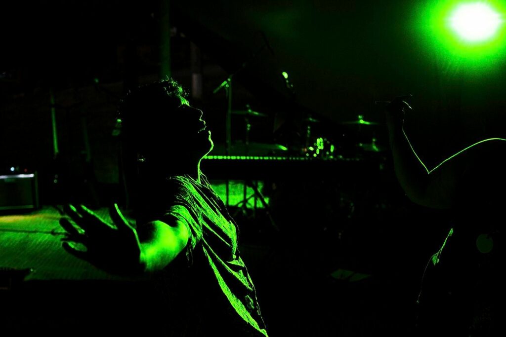A person raising their arms in a jubilant pose is illuminated by green stage lighting. In the background, musical instruments and equipment, including a drum kit and microphone stand, can be seen under the dim, dramatic lighting. Image by Documentary Wedding Photographer Lewis Bishop Photography.