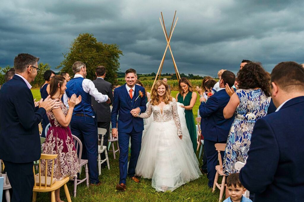 A bride and groom walk down the aisle outdoors, smiling and holding hands. Guests on either side of the aisle clap and celebrate. The groom wears a blue suit, and the bride wears a white dress with a tulle skirt. The sky is cloudy, and the background is lush and green. Image by Documentary Wedding Photographer Lewis Bishop Photography.