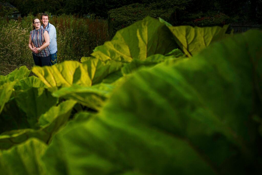 A couple stands closely together, smiling while embracing in a garden. They are surrounded by lush greenery and partially obscured by large, vibrant green leaves in the foreground. The atmosphere appears serene and natural. Image by Documentary Wedding Photographer Lewis Bishop Photography.