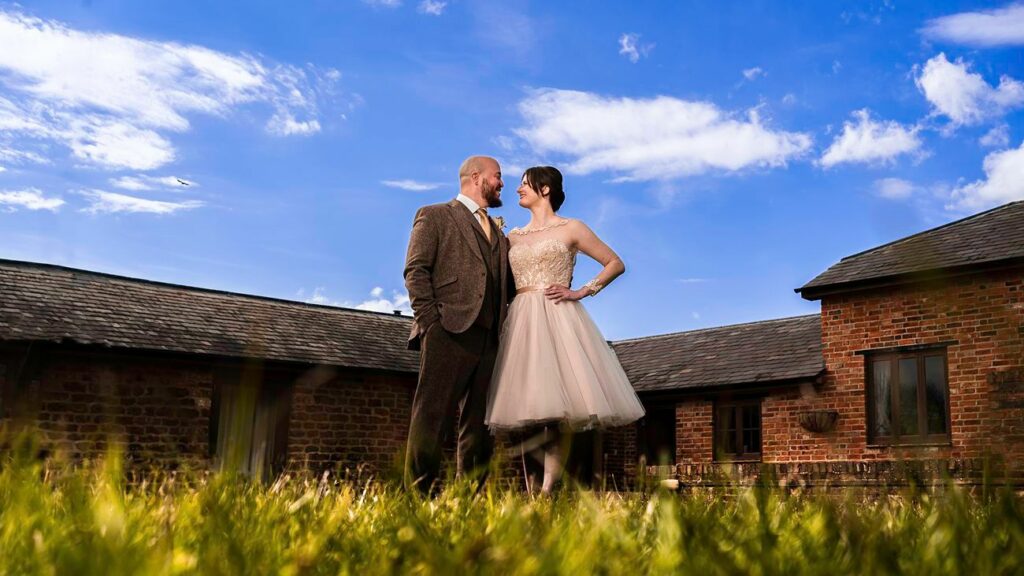 A couple stands together outside on a sunny day, smiling at each other. The man, in a brown suit, and the woman, in a beige dress, are in front of rustic brick buildings with slate roofs. The bright blue sky with scattered clouds is visible above them. Image by Documentary Wedding Photographer Lewis Bishop Photography.