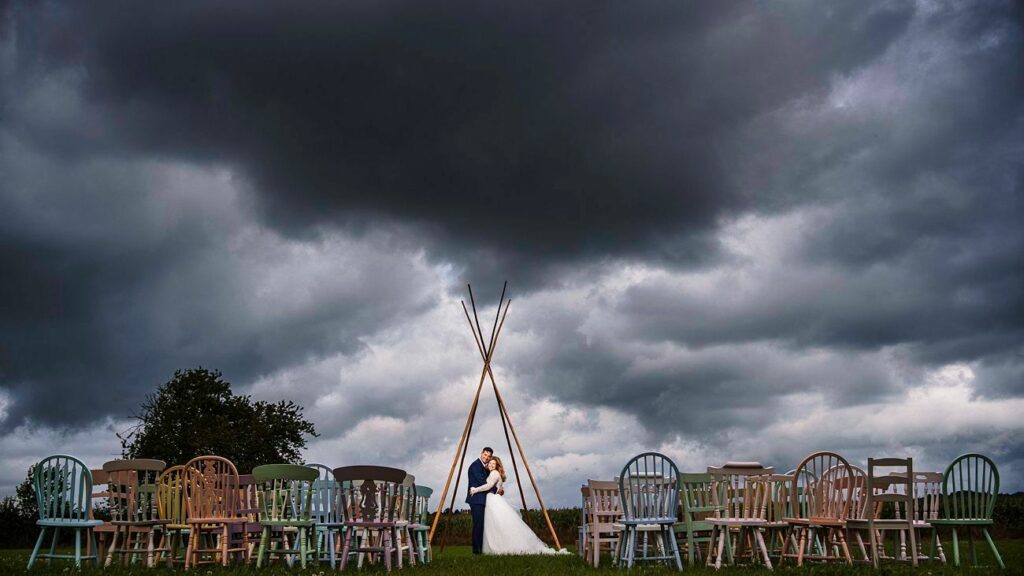 A newlywed couple embraces under a tipi-style wooden structure in an outdoor setting. They stand on green grass, and behind them, the sky is dark and stormy. The scene is framed by rows of colorful, assorted chairs arranged for a wedding ceremony. Image by Documentary Wedding Photographer Lewis Bishop Photography.