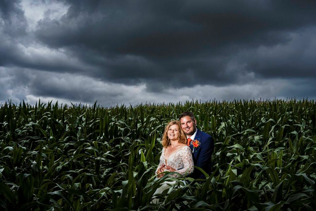 A bride and groom sit together in a lush green cornfield under a dramatic, cloudy sky. The bride is wearing a lace wedding dress, and the groom is in a dark suit with a red boutonniere. Both are smiling and looking towards the camera. Image by Documentary Wedding Photographer Lewis Bishop Photography.