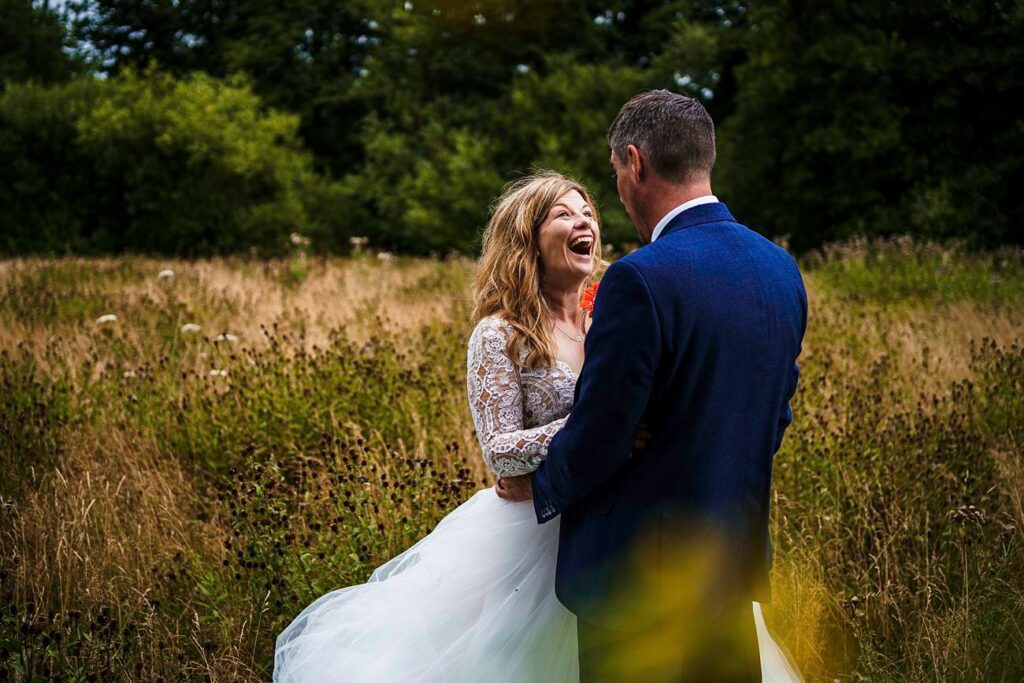 A laughing bride in a white wedding dress with lace sleeves and a groom in a blue suit stand in a wildflower meadow, surrounded by tall grass and trees. The bride has her hands on the groom's shoulders, and they appear to be sharing a joyous moment. Image by Documentary Wedding Photographer Lewis Bishop Photography.