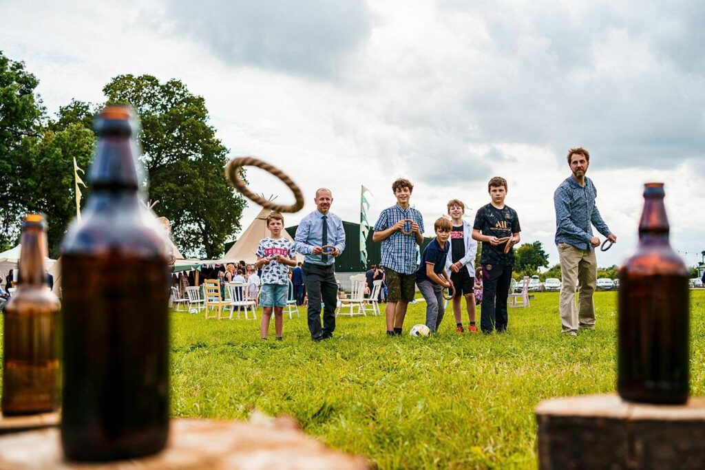 A group of six people, including children and adults, are playing an outdoor game at a grassy field event. They are attempting to throw rings over the tops of bottles. In the background are people sitting at tables under large green tents on a cloudy day. Image by Documentary Wedding Photographer Lewis Bishop Photography.
