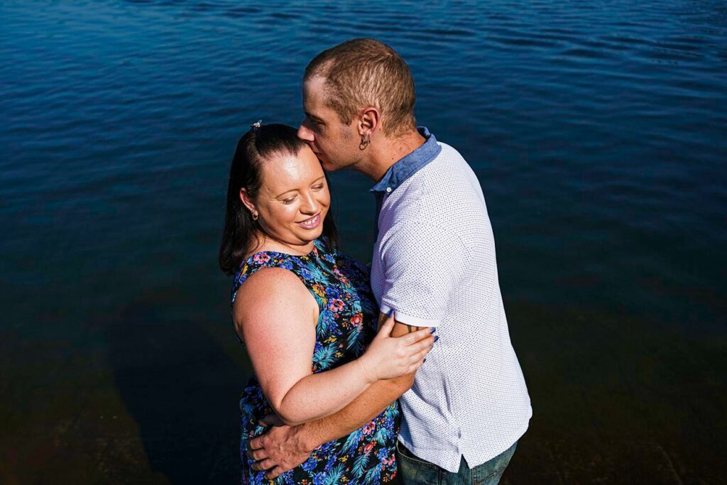 A couple stands close together by the water; the man, wearing a white shirt, kisses the forehead of the woman, who is wearing a blue floral dress, as she smiles and holds onto him. The background features calm, blue water. Image by Documentary Wedding Photographer Lewis Bishop Photography.