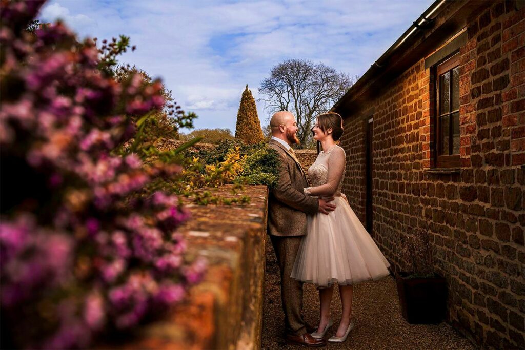 A couple stands close, gazing at each other, in a rustic outdoor setting with a brick wall and plants. The bride wears a knee-length dress, and the groom is in a suit. Pink flowers are in the foreground, and trees are visible in the background against a blue sky. Image by Documentary Wedding Photographer Lewis Bishop Photography.