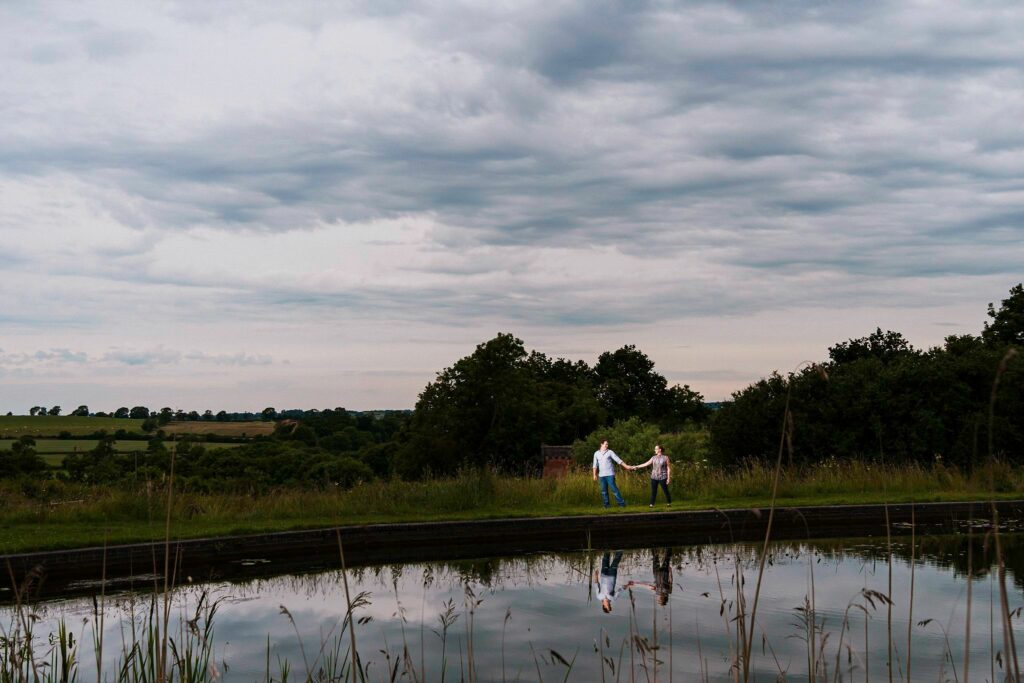 Two people hold hands while walking along a narrow path next to a reflective body of water, with an expansive cloudy sky and green landscape stretching into the distance. Tall grasses and trees frame the scene, creating a serene countryside setting. Image by Documentary Wedding Photographer Lewis Bishop Photography.