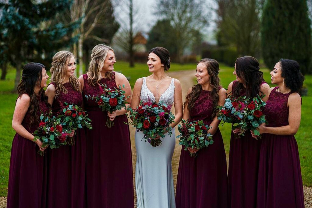 A bride in a white gown stands outside with five bridesmaids in matching burgundy dresses. Each bridesmaid holds a bouquet of red and green flowers, and the group smiles and looks at each other with a backdrop of trees and greenery. Image by Documentary Wedding Photographer Lewis Bishop Photography.