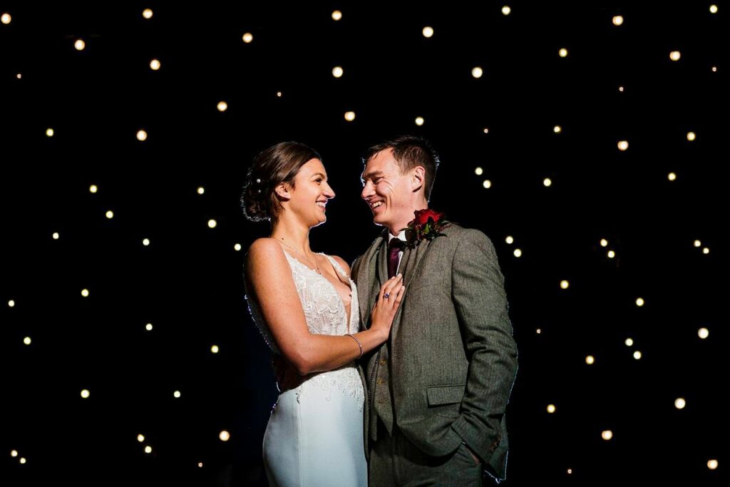 A bride in a white dress and a groom in a gray suit stand closely, smiling at each other against a backdrop of numerous small, glowing lights. The dark background enhances the warm, romantic ambiance created by the twinkling lights. Image by Documentary Wedding Photographer Lewis Bishop Photography.