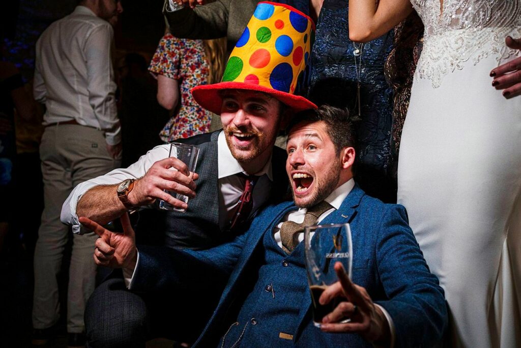 Two men in suits are posing at a festive event, one wearing a colorful polka dot top hat. They are holding drinks, pointing, and smiling enthusiastically. Other people, partially visible, are in the background, dressed in formal attire. Image by Documentary Wedding Photographer Lewis Bishop Photography.