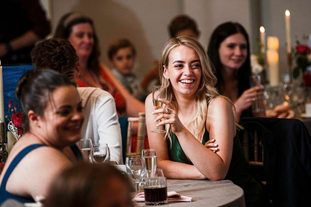 A smiling woman in a green dress holds a champagne flute while seated at a table during an event. Other guests, including adults and children, are seated nearby, engaged in conversation. The background shows lit candles and floral decorations on the tables. Image by Documentary Wedding Photographer Lewis Bishop Photography.
