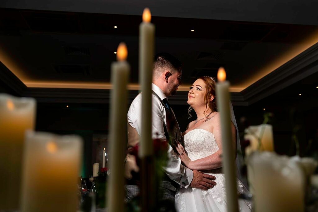 A bride and groom share an intimate moment, looking into each other's eyes while standing close together. They are surrounded by lit candles, creating a warm and romantic atmosphere. The bride wears a white gown, and the groom is dressed in a suit. Image by Documentary Wedding Photographer Lewis Bishop Photography.