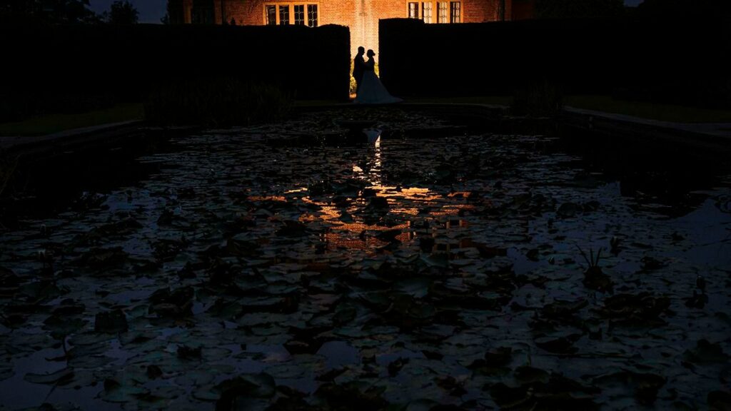 A silhouetted couple stands close together in front of an illuminated doorway of a brick building at night. The dark scene is enhanced by the reflection of the building's warm light on a pond filled with lilypads in the foreground. Image by Documentary Wedding Photographer Lewis Bishop Photography.