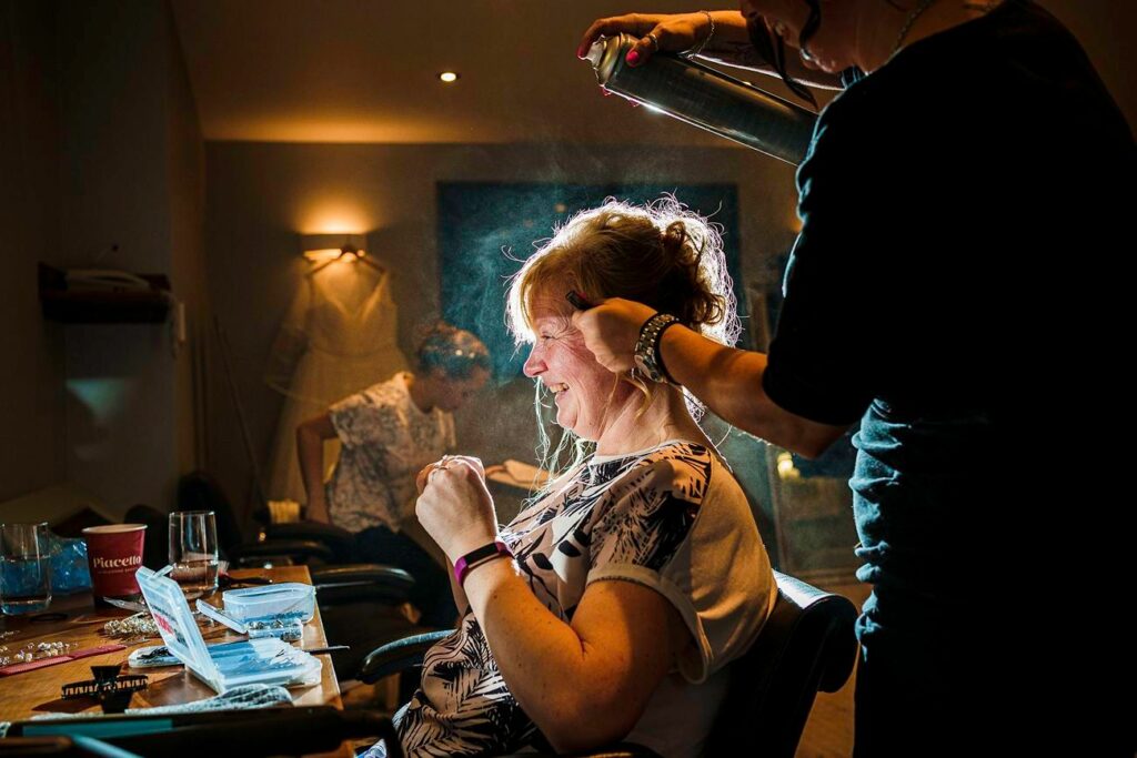 A woman with light hair sits in a chair, smiling as another person styles her hair. The stylist is seen spraying hairspray, and a wedding dress hangs in the background. Various makeup and hairstyling tools are scattered on the table in front of them. Image by Documentary Wedding Photographer Lewis Bishop Photography.