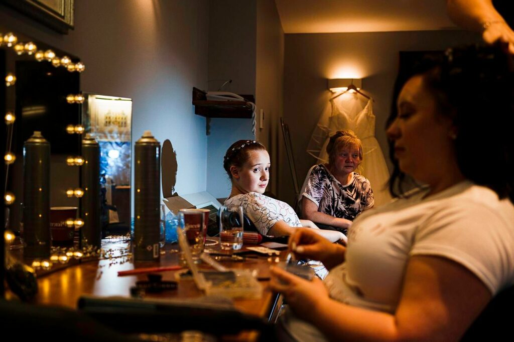 In a dimly lit room, a young girl with braided hair sits next to a woman getting her hair done. A mirror with lights, hair products, and beauty tools are on a cluttered table. Another woman, seated in the background, looks on. The scene appears calm and focused. Image by Documentary Wedding Photographer Lewis Bishop Photography.